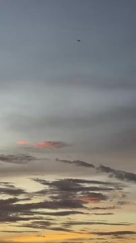 This media file depicts a serene sunset scene in Khan Mean Chey, Cambodia. The sky is filled with a mix of gray and dusky clouds, tinged with hints of orange and pink, suggesting the final moments of daylight. A lone airplane is visible as a small silhouette against the vast expanse of the sky, indicating travel or transit. The clouds are layered and textured, with some appearing wispy and others more dense, creating a dynamic visual. The overall impression is one of peacefulness and the transient beauty of nature. The time of day is clearly sunset, characterized by the soft, warm light and the atmospheric colors. No people are visible, and no text is present in the frame.