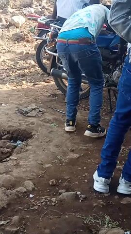 Two people, one wearing jeans and black and yellow shoes, and the other wearing blue jeans and white shoes, are standing on a dirt path near a motorcycle. The first person's rainbow-striped underwear is visible above their jeans. The motorcycle has a chrome frame and is parked on the left side of the frame. The dirt path is littered with small debris and scattered vegetation.