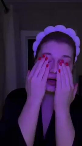 A woman with red nail polish wearing a black robe and white cloud-shaped headband is standing in a dim bathroom. Her face and hands are lit with purple light. She touches her face with both hands and makes hand gestures including pointing upwards and forming the letter L with her fingers. A white door is behind her on the right, and a white shower curtain is on the left. Music is playing in the background.