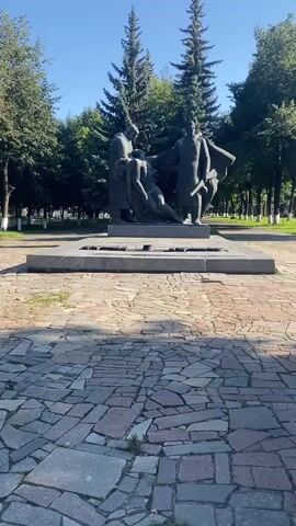 The video shows a statue in a park on a sunny day. The statue depicts three figures. One figure is lying down, seemingly injured or deceased, and is being held up by two other figures. The two figures appear to be men, and they are dressed in what looks like historical or military attire, with one holding a rifle. The statue is made of dark material, possibly bronze or stone. It is mounted on a large, rectangular stone base. The base is situated on a paved area made of irregularly shaped, colorful stones. Behind the statue, there are tall evergreen trees and deciduous trees with green foliage. The sky is clear and blue. The overall setting suggests a monument or memorial in a public space.