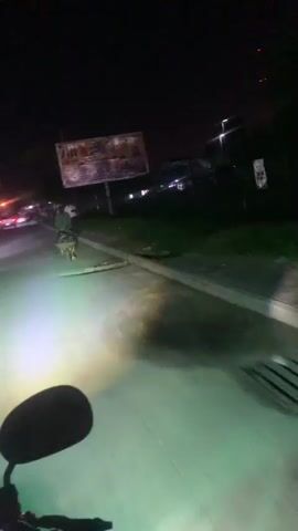 The video is taken from a first-person perspective, presumably from a motorcycle at night. The rider's hand and a rearview mirror are visible in the foreground. A bright headlight illuminates the road ahead, casting a green glow. To the left of the road, there is a grass verge and a concrete curb. Further back, a large billboard with text and images of people is visible. A person is seen standing near the billboard, holding something over their shoulder. In the distance, there are streetlights and trees, and some structures that appear to be buildings. The overall scene suggests a nighttime journey on a road with some urban or semi-urban features.
