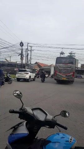 The video shows an outdoor scene, likely a street intersection in an urban area. The perspective is from the viewpoint of someone sitting on a parked scooter, with the scooter's handlebars and mirrors partially visible in the foreground. In the middle ground, a white SUV is turning through the intersection, while several motorcycles are parked on the left side of the frame. Two buses are visible, one parked on the left and one partially visible on the right, both labeled "Kairosbus.com". The background consists of various buildings, power lines, and utility poles creating a busy, somewhat cluttered visual.  A banner hangs on the left. The sky is overcast, suggesting a cloudy day.  Several people are present, some standing by the motorcycles, and others riding motorcycles through the intersection.