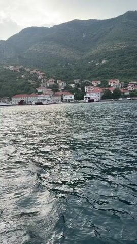 A vertical, full-shot video shows a body of water in the foreground with a village and a mountain in the background. The water is dark blue with sunlight reflecting off the waves, creating a rippling effect. The village is situated along the shore, with buildings featuring red-tiled roofs. The houses are nestled into the lush green mountainside. The sky is cloudy.