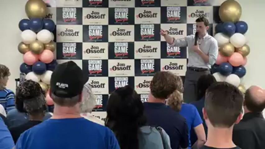 An indoor political event is underway, with a male speaker addressing an audience. The speaker, wearing a light-colored button-up shirt and gray trousers, stands on a small stage or raised area, holding a microphone and gesturing with his right hand. Behind him is a large checkered backdrop featuring the phrases "GROUND GAME" and "Jon Ossoff Georgia" repeated across its surface. Flanking the backdrop are two tall balloon columns, decorated with balloons in shades of blue, white, gold, and pink.

The audience, composed of diverse individuals, is seated or standing, facing the speaker. Many attendees are seen from behind, indicating the camera's position among the crowd. One person in the immediate foreground wears a black baseball cap with "ATLANTA UNITED" printed on the back. The overall atmosphere suggests an engaged and attentive audience at what appears to be a campaign rally or town hall meeting.
