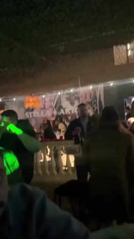The video appears to capture a lively indoor scene, possibly a bar or club, with numerous people present. The lighting is dim and dynamic, with various colored lights, including orange, blue, green, and red, flashing and illuminating the space. People are seen standing, talking, and some appear to be dancing or moving to music, though the audio is not provided for analysis. The environment looks somewhat enclosed, with a textured ceiling that resembles netting or camouflage. In the foreground, the faces of several individuals are partially visible, their expressions suggesting engagement with the environment. In the background, more people are discernible, some appearing to be socializing at tables or bars. The overall impression is one of a vibrant social gathering.