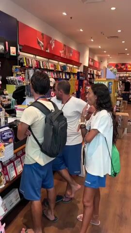 The video shows the interior of a bookstore. In the foreground, three people are visible. On the left, a person with dark hair, wearing a tan t-shirt and blue shorts, faces away from the camera, with a black backpack on their back. In the center, a man with a shaved head and a beard, wearing a white t-shirt and blue shorts, faces the camera and gestures with his left hand. To the right, a person with long, dark curly hair, wearing a white t-shirt and blue shorts, faces the camera and gestures with their left hand. Behind them, a checkout counter is staffed by two women. One woman, wearing a black t-shirt, stands behind the counter. Another woman, wearing an orange shirt, stands to her left and appears to be handing something to a customer. Bookshelves filled with books are visible in the background. Several displays of merchandise, including plush toys and stationery, are positioned near the checkout counter. The environment appears well-lit with overhead lighting.