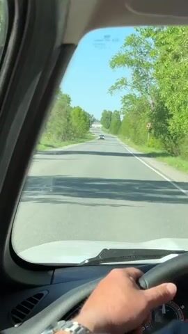 A driver, wearing a watch and a blue striped shirt, is driving a car on a two-lane road. The road is straight, with a slight rise and fall, and has a white car in the distance. Trees line both sides of the road. The sky is blue and clear. A road sign is visible on the right side of the road. The camera is inside the car, behind the steering wheel, showing a driver's point of view.