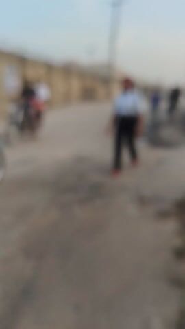 A blurred video, filmed from a low angle, shows a person standing near a building. The person has red-colored dreadlocks that are styled into several small buns. The person is wearing a light blue button-down shirt, dark pants, and a wristwatch. The building appears to be a multi-story structure with a staircase visible on the left side of the frame. The background is out of focus.