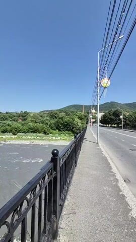 A clear, bright day with a blue sky over a bridge. The camera moves forward along a sidewalk next to a road. On the left side of the sidewalk is a black metal railing overlooking a river with a rocky bank. Trees and green hills are visible across the river. On the right side of the road, there are streetlights and power lines. A black car drives past in the distance. Beyond the road and trees, mountains are visible in the background.