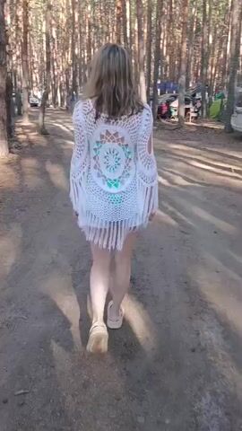 A woman with blonde hair walks away from the camera down a dirt path in a wooded area. She is wearing a white crocheted cover-up with fringe and blue bikini bottoms. The cover-up has a circular mandala-like design on the back. She is wearing tan platform sandals. The path is lined with tall, thin trees. In the background, there are some cars, tents, and other camping equipment. The sun is shining, casting shadows on the path.