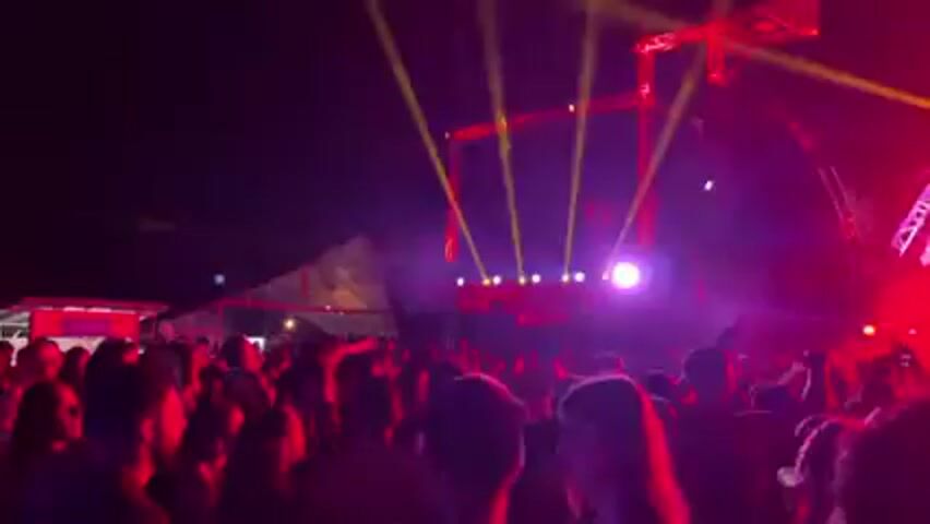 The video shows a concert or festival with a large crowd of people facing a brightly lit stage. The stage has a complex structure with multiple levels and illuminated by various lights, including yellow spotlights and red and white lights. The crowd is a mix of silhouettes and individual figures, suggesting a nighttime setting. The lighting creates a dynamic and energetic atmosphere.