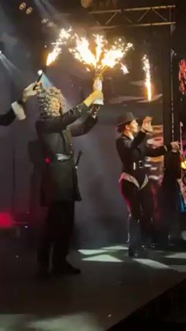 The video shows several people on a stage in a dim lighting environment. A person dressed in a dark suit and grey curly wig stands out, holding a bottle with sparklers shooting out the top. To the right, a woman in a black outfit and hat holds a lit sparkler in her right hand, performing an arm movement. Other individuals surround them, some holding lanterns, adding to the stage's theatrical feel. The background includes stage lights and abstract projections.