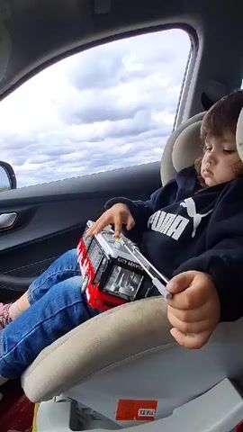 A young child sits in a car seat, wearing a black Puma hooded sweatshirt and blue jeans.  They hold a toy bus in their lap, manipulating a white lever on the bus. The bus is red and silver and has "CITY SERVICE" printed on the front. The car window shows a cloudy sky. The car's interior is dark, with a light-colored seat.  The child appears focused on playing with the toy bus.