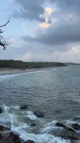 A video of a beach with rough waves crashing onto rocks in the foreground. In the background, a sandy beach meets a grassy hill dotted with small buildings. The sky is overcast with dark clouds.