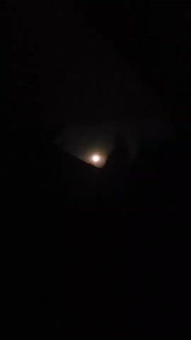 A bright, full moon is partially obscured by dark, silhouetted objects, possibly trees or mountains, in the foreground. The scene is dark, with the moonlight providing the primary illumination. The video is shot at night.