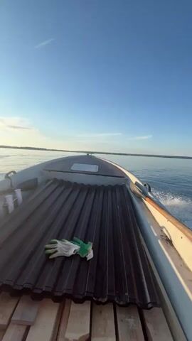 This is a POV shot from a boat moving across a body of water. The boat appears to be an inflatable raft with some wooden planks and what looks like metal sheeting in the foreground. A pair of green and white work gloves rests on the metal sheeting. A plastic water bottle is also visible on the wooden planks.

The scene is set on a calm lake or wide river under a clear blue sky with a few wispy clouds. The sun is shining brightly, casting a shimmering reflection on the water, indicating it is likely morning or late afternoon. A contrail from an airplane is visible high in the sky. The boat is creating a wake as it moves, with foamy water churning behind it. The distant shoreline is visible as a faint, tree-lined horizon. The overall mood is peaceful and serene, suggesting a leisurely boat trip or journey.

There is no visible text in the media file.