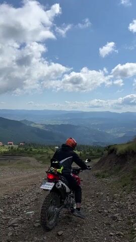 A person wearing a black jacket and an orange helmet sits on a dirt bike on a rocky, uphill path. The dirt bike is facing away from the camera. The person is wearing black pants and gray shoes. In the background, there are rolling hills covered in green trees and some buildings. The sky is blue with white, fluffy clouds. The scene appears to be a mountainous landscape on a sunny day.