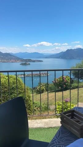The video pans across a scenic view of a large body of water, likely a lake, with mountains in the distance. In the foreground, there is a black metal railing, with green foliage and some pink flowers visible behind it. On the right side of the frame, a wooden box with some items inside is placed on a patterned surface. The water itself is blue and calm, with several small islands dotting its surface. Boats are visible on the water, some of which appear to be moving. The sky is bright blue with a few white clouds. The scene appears to be from a balcony or terrace, suggesting a viewpoint overlooking the landscape.