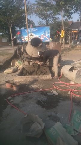 This media file captures an outdoor scene in Gebangilir, Indonesia, where a person is actively engaged in mixing cement. The individual, wearing a dark long-sleeved shirt, light-colored pants, and a white cap, is using a shovel to work with wet cement in a rectangular trough. Behind them, a large concrete mixer drum is positioned, suggesting it has recently been used or is about to be used further. Several bags of cement are visible in the foreground and to the left of the mixer, some marked with "50," indicating their weight. Red hoses snake across the ground, likely for water supply. The setting appears to be a construction or renovation site, with a dusty, uneven ground surface and scattered building materials. In the background, trees with sparse foliage line a pathway, and a building with a tiled roof is visible. A large blue banner is stretched across a pole, displaying text and an emblem. The lighting suggests it is daytime, possibly late morning or early afternoon, with the sun casting shadows. The overall atmosphere is one of work and industry. The visible text on the banner, though partially obscured, includes Indonesian words like "PELATIHAN" (training) and "ARMY," hinting at a possible connection to a military or vocational training event. The presence of multiple concrete mixers and bags of cement points to a significant construction or repair project underway.