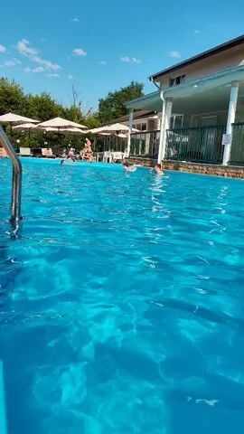 The video captures a sunny day at an outdoor swimming pool. In the foreground, the bright blue water of the pool is visible, with ripples and light reflections. A metal ladder is present on the left side of the frame. Further in the pool, several people are swimming and interacting. One person is seen on the far left, partially obscured by the ladder. Closer to the center, a child wearing a flotation device is in the water, appearing to swim. Another person is to their right, also in the pool. On the right side of the pool, two more individuals are swimming, one of whom appears to be gesturing. In the background, a building with a light-colored facade and a covered patio area with white umbrellas and tables is visible. Trees and greenery surround the area. The sky is a clear blue with a few white clouds. The overall atmosphere suggests a leisurely and recreational setting.
