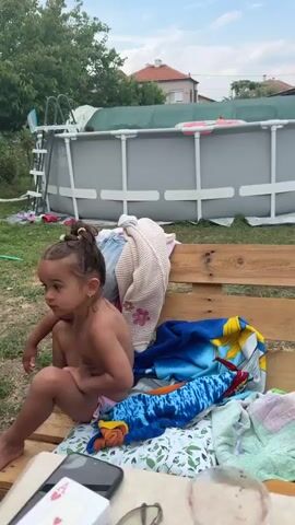 This video features a young girl sitting on a wooden bench outdoors, likely in a backyard. She is wearing a swimsuit, and her hair is wet and pulled up in a bun, suggesting she has recently been in the water. She appears to be playing or interacting with someone off-camera, her expressions changing from a pensive look to excitement and open-mouthed joy as she gestures with her hands.

In the background, an above-ground swimming pool is visible, complete with a ladder, further confirming a pool-related activity. The setting appears to be a grassy yard with some greenery and a house in the distance, indicating a residential area. The sky suggests it is daytime, likely a warm day given the pool activity. The girl's emotions are a central focus, transitioning from calm observation to animated engagement. There is no visible text in the video. The context provided indicates the location is Svilengrad, Bulgaria.