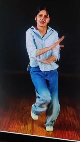 A young girl with dark hair styled in pigtails, wearing a light blue collared shirt and loose-fitting blue jeans, dances in front of a dark background. The floor is made of polished wood. She moves her arms and torso with a lively rhythm, her facial expression animated. In one frame, she points upwards with her right hand.