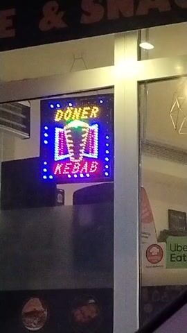 The video shows the exterior of a restaurant at night. A lit-up LED sign is visible in the window, advertising "DONER KEBAB". The sign is multicolored, with yellow lettering for "DONER", a green and white image of a kebab, and red lettering for "KEBAB". Blue LED lights surround the sign. The window also has a sticker advertising Uber Eats. Parts of the restaurant's name, "E & SNAC", are visible above the window.