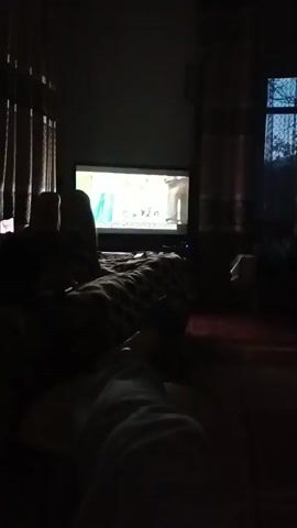 A person is lying on a bed, with their legs visible in the foreground, under a blanket. They are watching a television screen, which is displaying an advertisement for a clothing store. The advertisement features several young people in athletic wear, with the text "APPROX. 70%" superimposed on the screen. The room is dimly lit, with curtains on either side of the television. The time stamp on the video is 00:00 to 00:04.