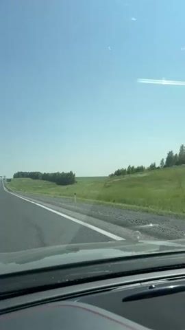 The video shows a first-person view from inside a car, looking out the windshield at a clear blue sky and a rural landscape. A paved road with a white line runs alongside the car. To the right of the road, there is a grassy field with scattered trees and bushes. The sun is shining, creating some glare on the windshield. The overall impression is of a scenic drive through the countryside on a sunny day.