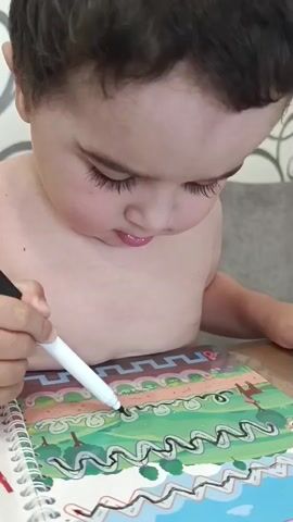 A close-up shot captures a young child with dark hair and thick eyelashes, looking down intently at a coloring book. The child is holding a white marker with a black tip in their right hand, coloring a black zigzag line within a green and brown wavy pattern. The child's bare chest is visible. The coloring book features images of trees and possibly a landscape. The background is out of focus but appears to be a wall with a textured pattern and a tabletop. The lighting is bright, suggesting an indoor setting. The child's mouth is slightly open, showing a hint of pink lips. The focus is on the child's concentrated expression and the act of coloring.