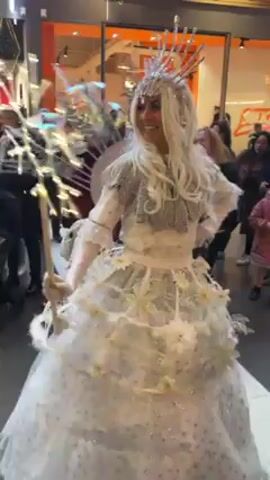 The video shows a group of performers dressed in elaborate, silvery white costumes walking through a shopping mall. One performer is wearing a long, flowing white dress with sparkling details, while another is wearing a short white costume with white fur leg warmers and large white wings adorned with lights. The third performer, positioned between the two, is elevated on stilts and covered in shiny silver materials, including a disco ball-like headpiece. People are standing around watching the performance, including a person pushing a baby stroller. The mall is decorated with large snowflake ornaments hanging from the ceiling.