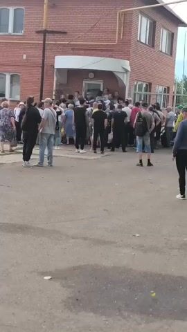 A crowd of people, dressed in casual clothing, are gathered outside a red brick building. The building has a white awning over a doorway. Some people are standing in line, while others are milling about. A man in a green shirt and khaki pants is walking away from the camera, towards the crowd. The ground is paved with asphalt. The weather appears to be overcast.