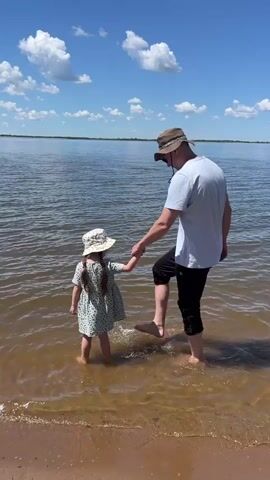 A man and a young girl are standing in shallow water near a sandy shore. The man, wearing a white t-shirt, black shorts, and a brown hat, holds the girl's hand. The girl, dressed in a light-colored floral dress and a matching hat, stands facing away from the camera. The man lifts his right leg, splashing water. The background shows a large body of water under a blue sky with scattered white clouds.