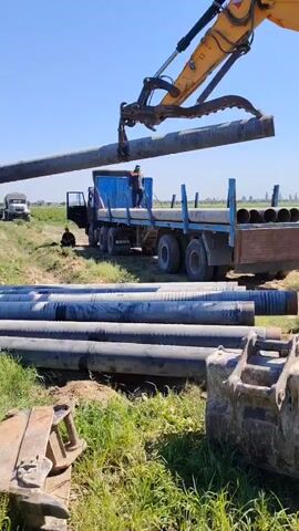 The video shows an excavator with a hydraulic grapple arm lifting a large pipe from a truck bed. The truck is blue and has a flatbed trailer. The pipe is being placed onto a stack of similar pipes lying on the ground. Several other pipes are already on the ground in the foreground, as well as a piece of excavator equipment. In the background, there is a white truck, a person sitting on the ground next to the truck, and a field of crops under a clear blue sky. The overall scene appears to be an outdoor industrial or construction site.