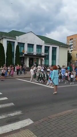 This is a vertical, outdoor video taken on a cloudy day. A group of children in uniforms, possibly for a school or parade, are walking across a street. They are accompanied by adults. In the background, there is a white building with a green roof and the sign "Прелагропромбанк" in Cyrillic. People are standing on the sidewalk watching the procession. There is a crosswalk visible in the foreground.