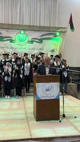 A graduation ceremony is in progress. A man in a suit stands behind a podium with a logo on it, smiling and looking toward the right. Behind him, a group of graduates in academic regalia are lined up. The graduates are wearing black gowns with silver trim and academic caps. Many of them are holding small Palestinian flags. The backdrop features a banner with Arabic text and the Palestinian flag. Lights are illuminating the stage. The graduates are clapping.