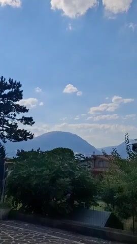 The media file captures a serene natural landscape with a towering mountain as the focal point. The sky is a vibrant blue, dotted with fluffy white clouds, suggesting a pleasant day. In the distance, the mountain's peak is gently rounded, its slopes covered in a soft, bluish-green hue, hinting at vegetation. A small, dark silhouette, possibly a paraglider, is visible against the vast sky, adding a touch of human activity to the otherwise natural scene.

In the foreground, lush green trees and foliage create a natural frame, with a few residential buildings peeking through the greenery. The architecture of the buildings suggests a European setting, with terracotta-tiled roofs. A stone-paved area is visible in the immediate foreground, indicating a man-made path or patio.

The time of day appears to be daytime, likely late morning or early afternoon, given the bright sunlight casting shadows and illuminating the scene. The weather is fair and clear, ideal for outdoor activities like paragliding. The overall mood is one of tranquility and natural beauty, with a subtle element of adventure introduced by the paraglider. The mention of Camerino, Italy, provides geographical context, placing the scene in a region known for its picturesque mountains and countryside. No visible text is present in the media file.