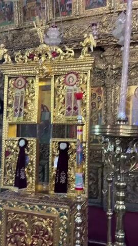 The video shows the interior of a church, focusing on an ornate iconostasis. The iconostasis is covered in gold leaf and features numerous religious icons depicted in a traditional Byzantine style. These icons portray various saints and biblical scenes. The video pans upwards to reveal a fresco on the ceiling, depicting Jesus Christ in the center, surrounded by angels. In the foreground, a tall, elaborately decorated candle stand is visible, with a candle lit on top. The lighting is dim, with golden reflections from the iconostasis illuminating the space. The overall atmosphere is one of solemnity and reverence.