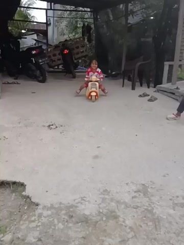A young girl with dark hair, wearing a red and white t-shirt with images and a red patterned skirt, is sitting on a small, cream-colored toy scooter. She is smiling and appears to be playing outdoors on a paved surface with patches of grass. In the background, there are motorcycles, wooden structures, and some chairs. The girl is seen pushing off the ground with her feet to move the scooter and at one point raises her fist in the air. She then turns her head and appears to wave.