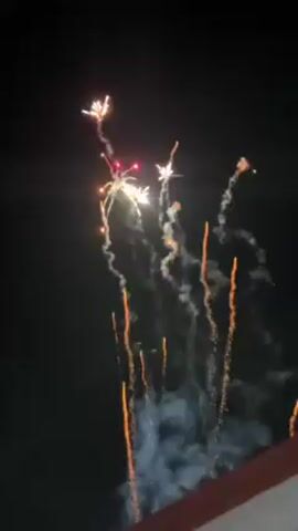 The video shows fireworks exploding in the night sky. The fireworks are a variety of colors, including red, white, and gold, and leave trails of smoke as they fall. The fireworks explode in different patterns, some resembling stars and others resembling palm trees. The bottom portion of the video shows a structure that seems to be the edge of a building, likely a porch or patio.