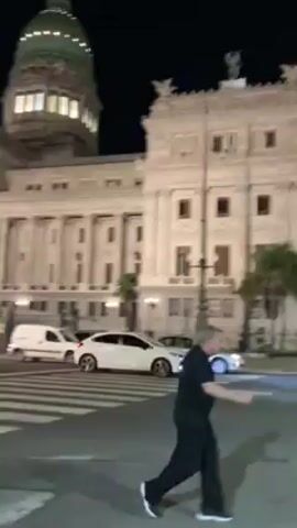 The video shows a night scene in a city with a large, illuminated building, possibly a government building or monument. People are crossing the street and walking around. There is traffic, including cars and a bus. In one area, there's a small gathering of people around what appears to be a makeshift memorial or display, possibly related to a protest or demonstration. Some people are taking photos. The lighting is a mix of streetlights, building illumination, and vehicle headlights.