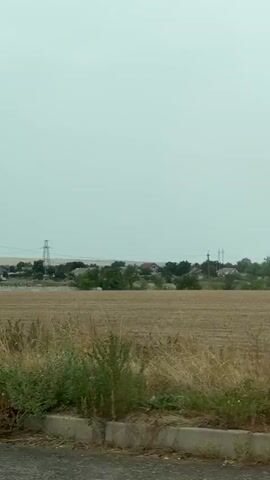 The video shows a rural landscape with fields, trees, and buildings. There are power lines and transmission towers visible in the distance. In the foreground, there is a concrete structure and vegetation. The sky is overcast. The video appears to be shot from a moving vehicle.