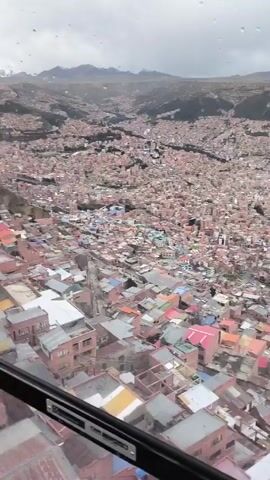 The video shows an aerial view of a densely populated city. The city appears to be situated in a valley, surrounded by hills or mountains in the background. The sky is overcast and grey. The buildings are mostly low-rise and built closely together, creating a uniform appearance. The rooftops are a mix of colors, including red, orange, grey, and blue. A few streets are visible snaking through the city. The viewpoint seems to be from a high vantage point, possibly a cable car or an elevated structure. The window frame of the vehicle from which the video is being taken is visible in the foreground. There are small droplets of water on the glass, indicating it may be raining or have recently rained.