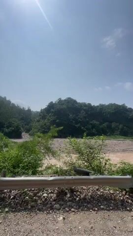 The video shows a landscape scene with a riverbed and lush green hills. The sun is shining brightly in the sky, casting a glare. The river appears to be shallow with a muddy bottom, and there are scattered rocks and vegetation along its banks. The hills in the background are covered with dense trees. The camera is panning slightly to the right, capturing the movement of the scene.