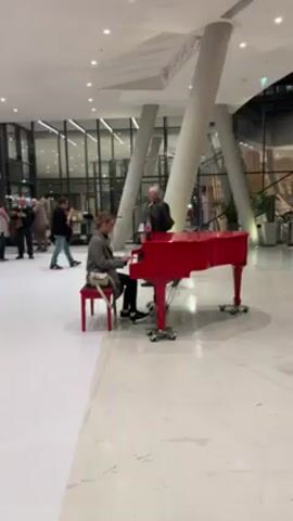 The video shows a woman playing a bright red piano in a large, modern building. She is seated on a matching red piano bench, dressed in a gray coat, black pants, and black shoes, with a small beige purse across her body. She wears glasses and has her hair pulled back. The piano is on wheels.  A man in glasses and a black jacket stands behind the piano, recording her with his phone. A bottle of Coca-Cola sits on top of the piano. Several people are walking around in the background. The building has tall, white support columns and large glass windows that reflect the scene inside. The floor is white with a faint checkerboard pattern.