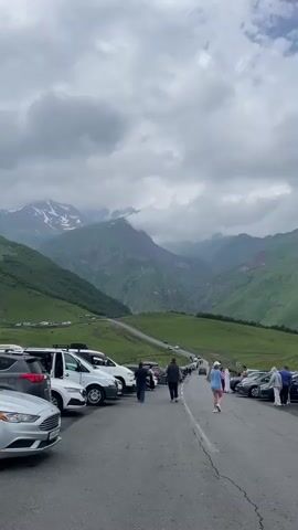The video shows a parking lot next to a winding road with a mountainous landscape in the background. Several cars, including white vans and SUVs, are parked along the side of the road. People are walking in both directions on the road and in the parking lot. The mountains are covered in green vegetation, and there are some patches of snow on the higher peaks. The sky is cloudy.