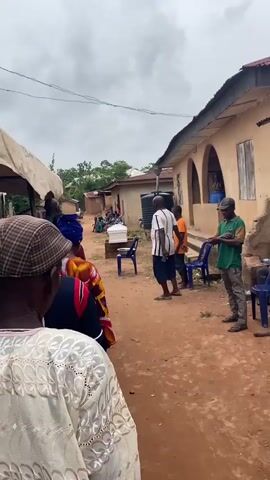 The video depicts an outdoor gathering in what appears to be a village. In the foreground, several individuals are seen from behind, facing away from the camera. The central focus is a white coffin placed on a raised platform in a dirt pathway. A blue plastic chair with a metal bowl on it sits in front of the coffin.

To the right of the coffin, a man wearing a white and black striped shirt and dark shorts stands, gesturing with his hands as if speaking. Behind him, a person in an orange shirt and another in a green shirt stand. A large black water tank is visible behind them.

Further into the scene, several people, predominantly women, are seated on the ground and on what look like benches in front of a row of buildings. The buildings have corrugated metal roofs and are constructed with earth-toned materials. The environment is rural, with vegetation visible in the background. The sky is overcast. The overall atmosphere suggests a somber event, likely a funeral or wake.
