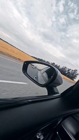 The video displays a first-person perspective of traveling in a vehicle. The camera is positioned inside the car, looking out the side window towards a side-view mirror. The mirror reflects the road ahead and other vehicles. The car is on a paved road, with a field of yellow crops on the left and trees on the right. The sky is cloudy with patches of blue. The overall impression is that of a scenic drive through the countryside.