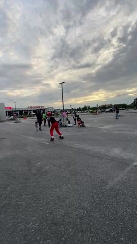 A man is roller skating on a paved lot. He is wearing a black tank top, red pants, a red helmet, and wrist guards. He is facing the camera and skating forward. The sky is cloudy. In the background, there are trees, buildings, and cars. Other people are visible in the background.