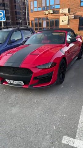 This is a video of a red convertible Ford Mustang parked in an outdoor parking lot. The car has black racing stripes on the hood and down the side. The car's black rims and red brake calipers are visible. The car is parked in front of a brick building with several windows and air conditioning units. A blue SUV is parked to the left of the Mustang. A sign on the building reads "147" and has some Cyrillic text. The sky is visible in the background. The lighting suggests it is daytime, likely late afternoon or early evening, with the sun casting soft shadows. The scene appears to be in a relatively urban or suburban area, possibly an apartment complex or commercial building. The video focuses on the exterior of the car, with a slight camera pan to the right. There is no visible activity or people in the video.